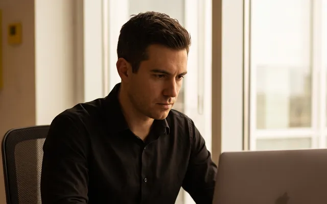 A founder working at a laptop with camera equipment, printed photo layouts and a yellow notebook on the desk, drafting content by hand.